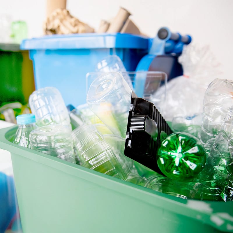 Volle Mülltonne mit recycelten Plastik Flaschen. Im Hintergrund ist eine Blaue Tonne für das recyclen von Papier und eine Gelbe Tonne für das recyclen für Verpackungen zu sehen.